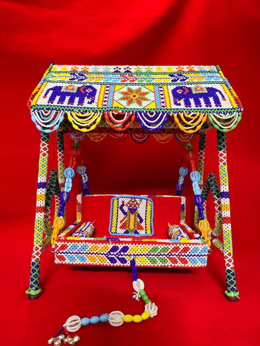 Miniature beaded swing with four decorated pillars, a canopy featuring elephant motifs, a beaded seat, and colourful hanging ornaments
